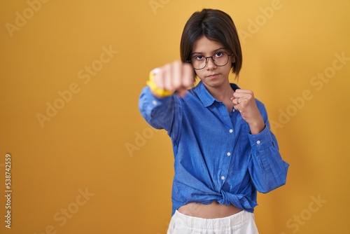 Young girl standing over yellow background punching fist to fight, aggressive and angry attack, threat and violence