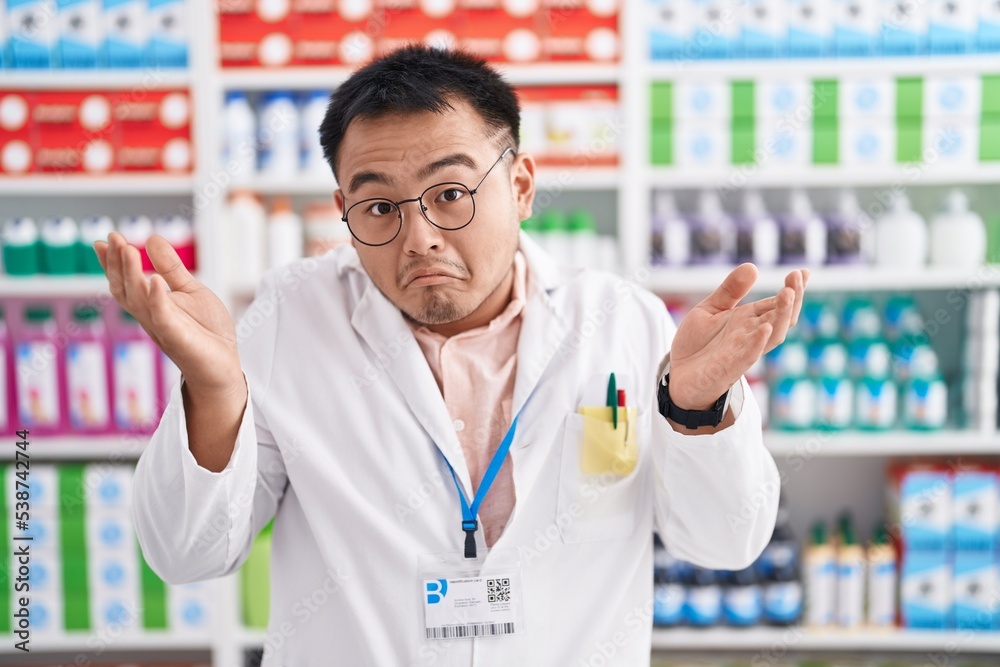 Chinese young man working at pharmacy drugstore clueless and confused expression with arms and hands raised. doubt concept. Chinese young man working at pharmacy drugstore clueless and confused expression with arms and hands raised. doubt concept.