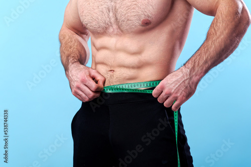 Strong bodybuilder measuring the width of his waist, putting a measuring tape over his abdomen, blue studio background