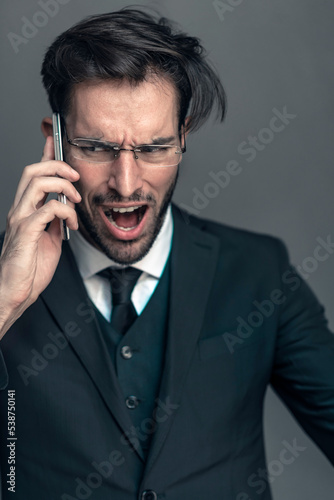 Angry and furious businessman shouting on his cell phone, studio portrait, grey background