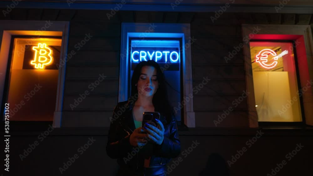 Young female crypto investor uses her smartphone buy or sell cryptocurrency at night against bright neon signs BITCOIN, CRYPTO and EURO on windows