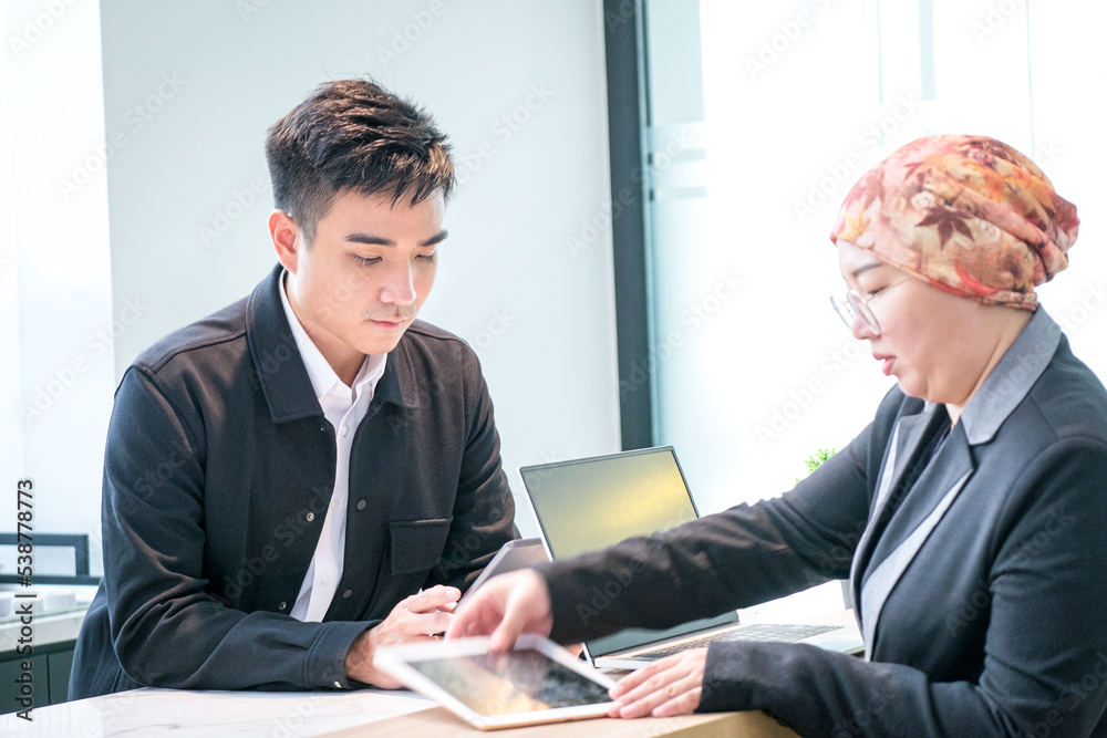 Fototapeta premium Two Asian business people working together, looking at digital tablets in office.