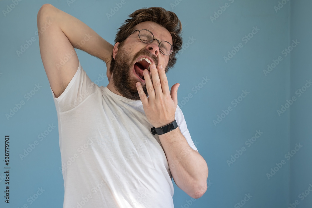 Bearded man in glasses and a white t-shirt yawns with mouth wide open, covering it with hand, he is sleepy or bored. Blue background. Bearded man in glasses and a white t-shirt yawns with mouth wide open, covering it with hand, he is sleepy or bored. Blue background.