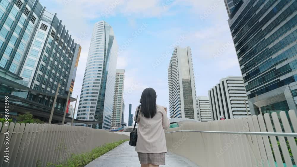 Back view of a businesswoman wearing a suit talking on the smartphone and holding a coffee cup walking on the street in the city center Back view of a businesswoman wearing a suit talking on the smartphone and holding a coffee cup walking on the street in the city center