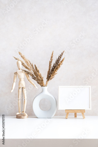 Empty picture frame mock up in minimalist interior with dried plant in vase on white table.