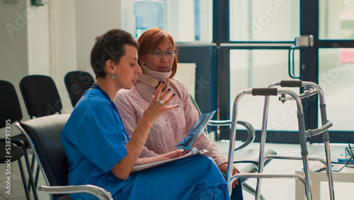 Asian woman with cervical collar looking at x ray scan results and receiving support from nurse. Assistant explaining radiography diagnosis to injured patient wearing neck foam brace.