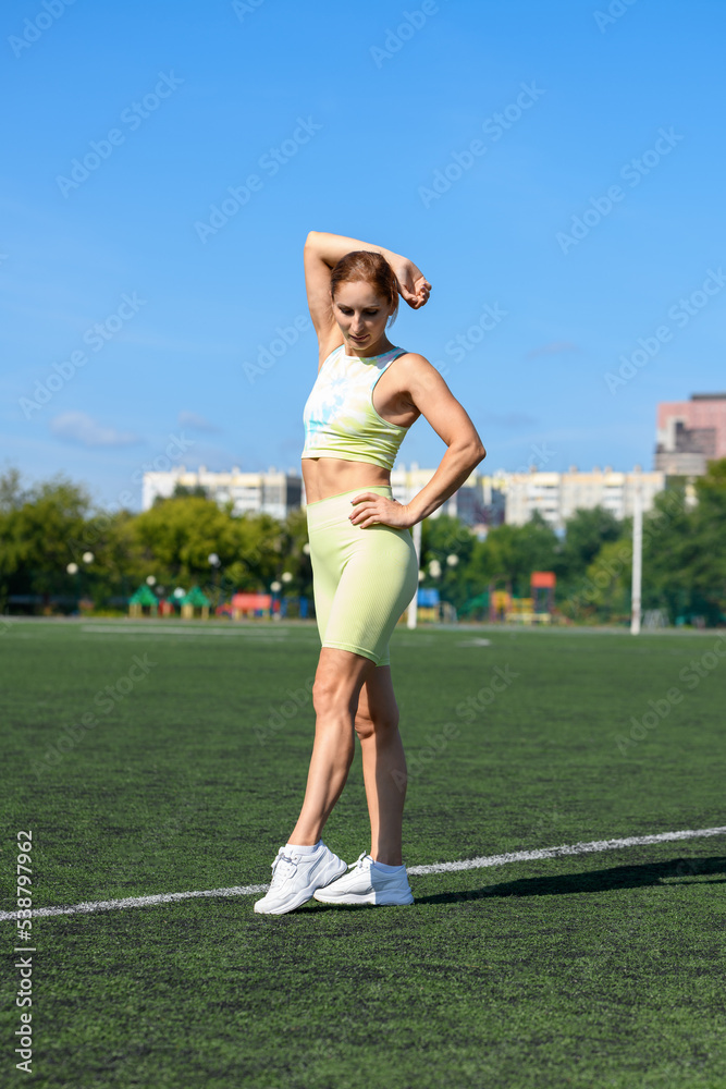 Women and sport. Smiling girl in summer sportswear stands on the grass stadium on a sunny day against the blue sky. Middle aged sportswoman dressed in sportsclothes
