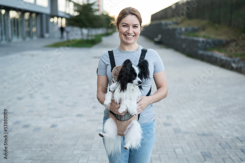 Happy caucasian woman walking with a dog in a backpack. Papillon Spaniel Continental in a sling.