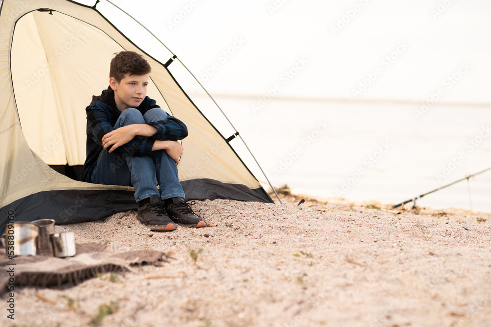 Family on the camping. Happy teenager is sitting near the tent at the beach. Caucasian boy having dreaming. Active male model. Travel, vacations and adventure.