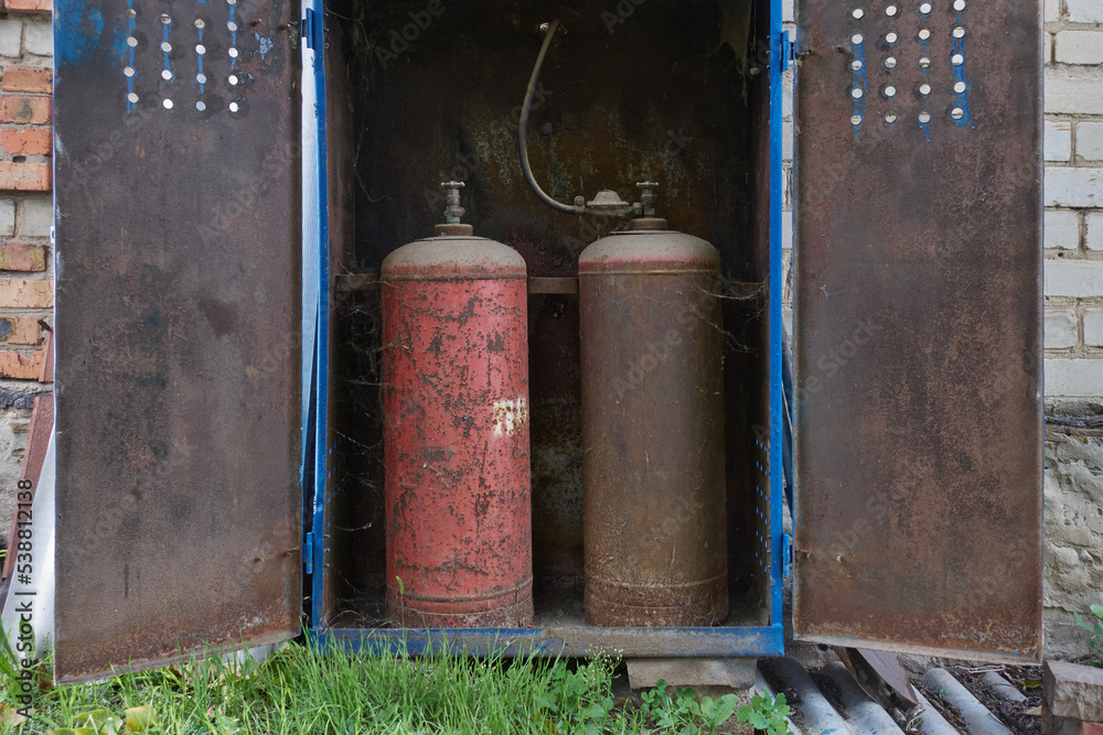 Old gas bottles. Rusty, cobwebbed, and dusty gas storage tanks sit in a metal storage box. There is a gas reducer on the cylinder. The rubber hose goes to the wall. Old gas bottles. Rusty, cobwebbed, and dusty gas storage tanks sit in a metal storage box. There is a gas reducer on the cylinder. The rubber hose goes to the wall.