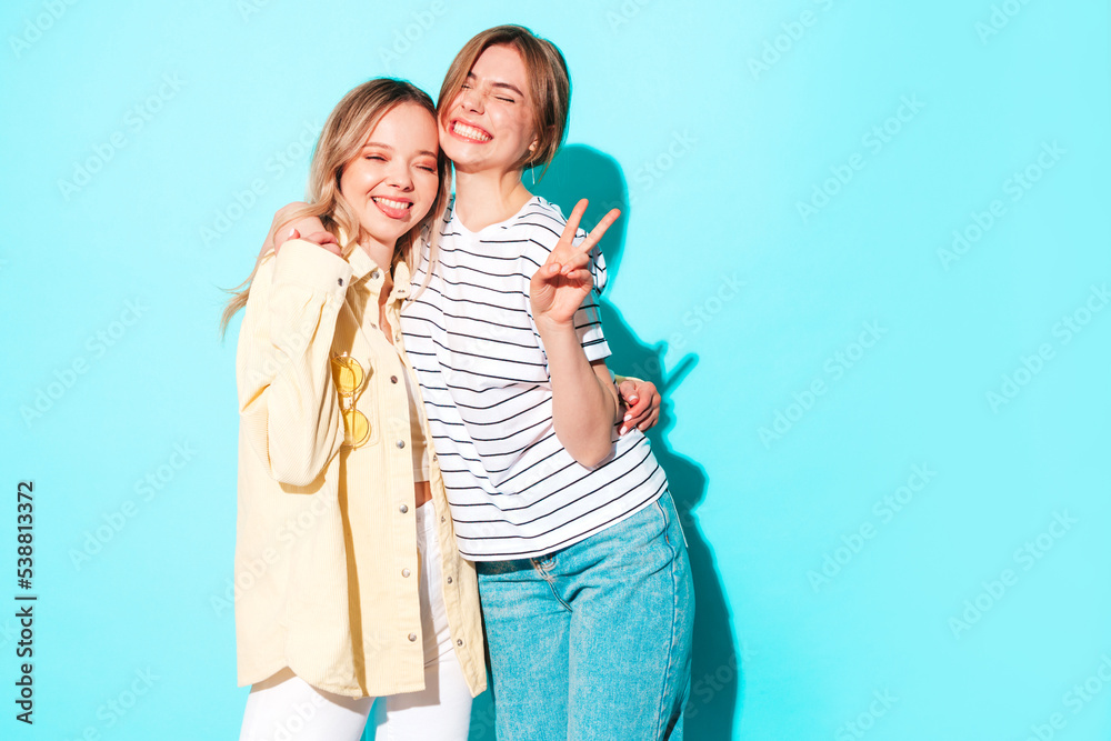 Two young beautiful smiling brunette hipster female in trendy summer clothes. Sexy carefree women posing near blue wall. Positive models having fun. Cheerful and happy