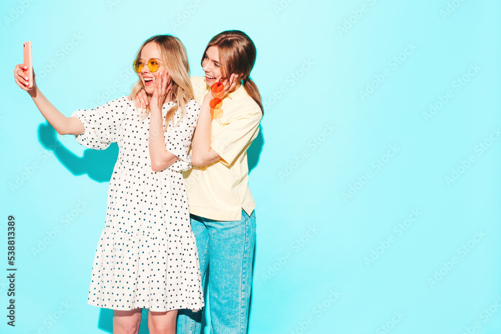 Two young beautiful smiling brunette hipster female in trendy summer clothes. Sexy carefree women posing near blue wall. Positive models having fun. Cheerful and happy