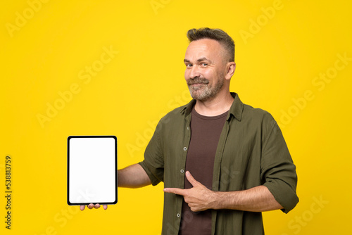 smiling handsome bristle with mustach mature man in green shirt pointing with finger and thumb up to his tablet pc with white mock-up screen holding in hand over yellow background