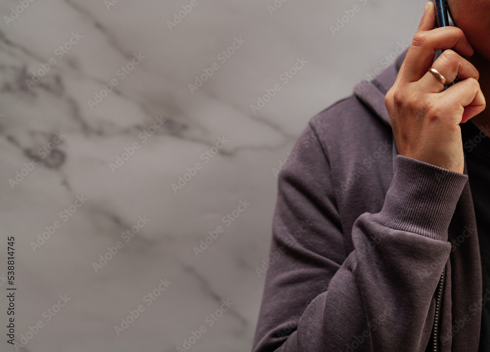 A woman in a sweatshirt holding a phone to her ear on a marble background. Place for text. Focus on the hand.