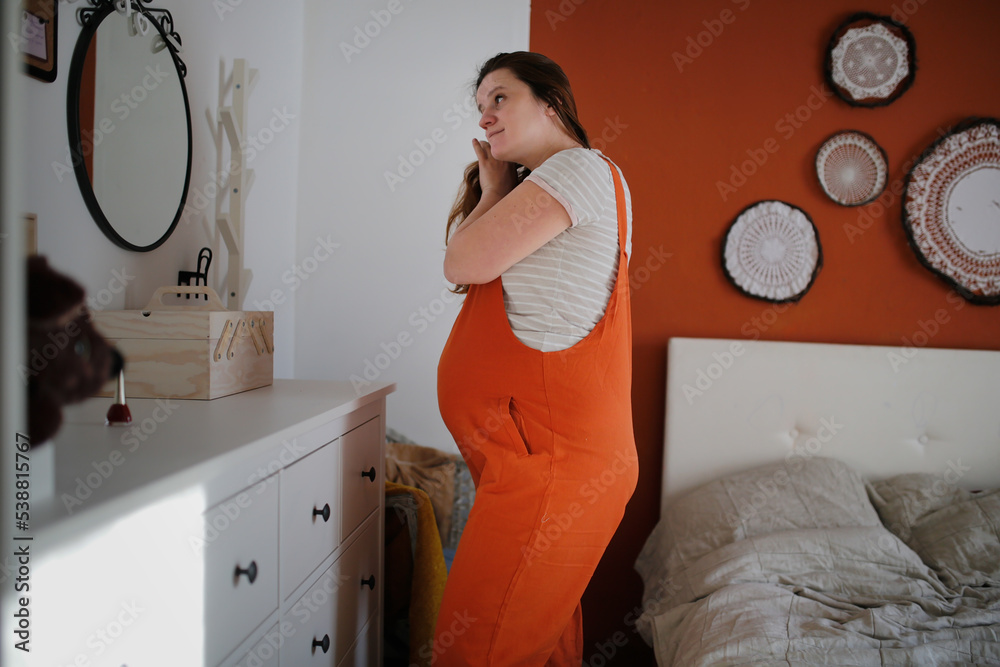 Pregnant plus size middle-aged woman combing her hair in front of mirror in bedroom.