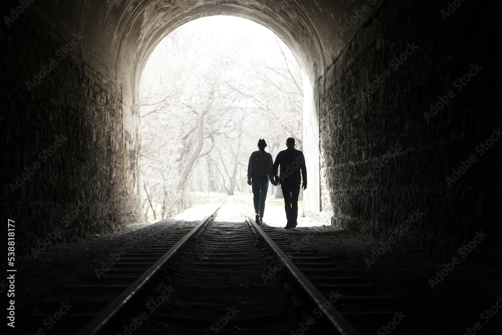 Couple walking hand in hand along the track through a railway tunnel towards the bright light at the other end, they appear as silhouettes against the light. back view.