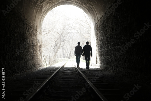 Couple walking hand in hand along the track through a railway tunnel towards the bright light at the other end  they appear as silhouettes against the light. back view.