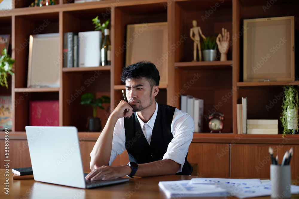 A portrait of a good-looking and discreet Asian man sitting at his desk with a thoughtful look on his computer A portrait of a good-looking and discreet Asian man sitting at his desk with a thoughtful look on his computer