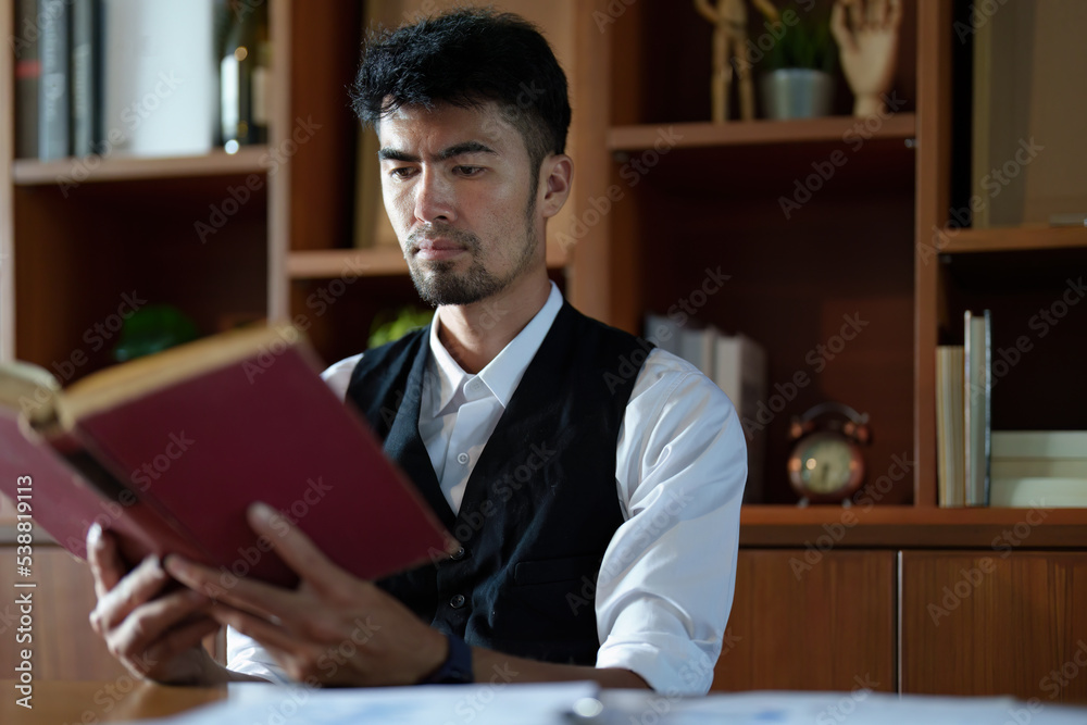 A portrait of a handsome and discreet Asian man sitting at his desk reading a book A portrait of a handsome and discreet Asian man sitting at his desk reading a book