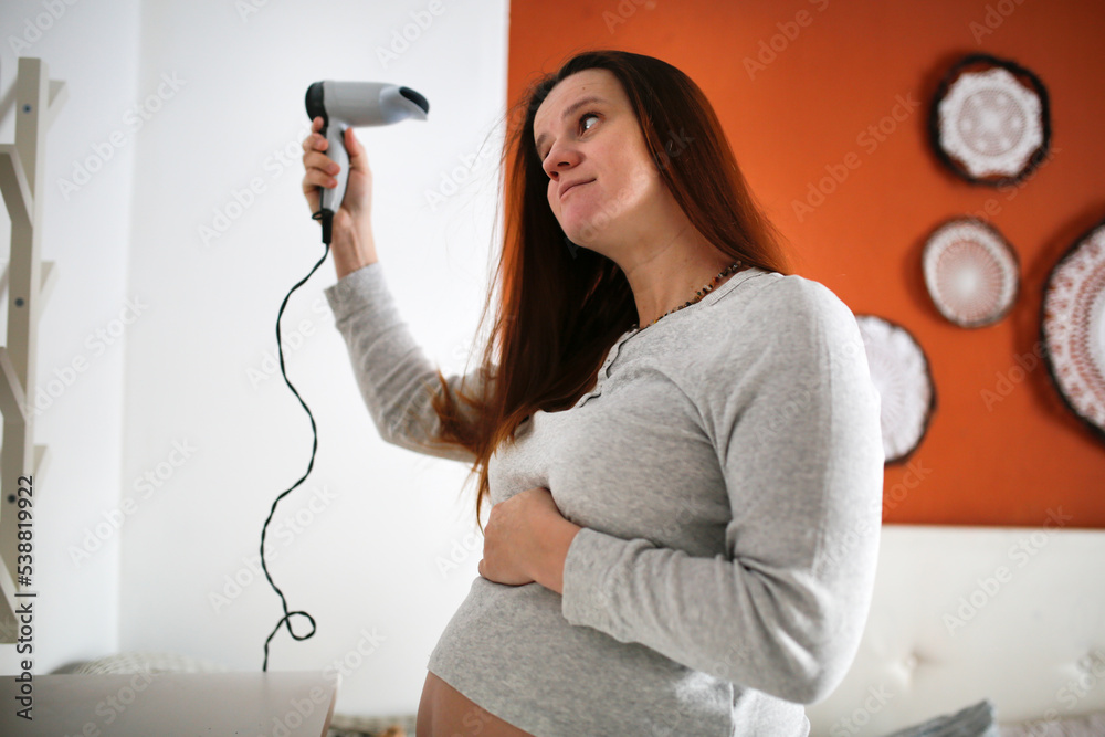 Pregnant woman in pajamas dries her hair with hair dryer in bedroom. Pregnant woman in pajamas dries her hair with hair dryer in bedroom.