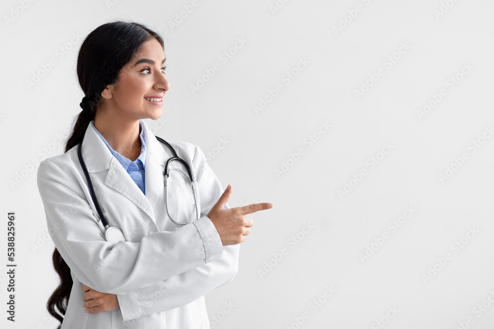 Smiling millennial indian woman doctor, family therapist in white coat show finger an empty space Smiling millennial indian woman doctor, family therapist in white coat show finger an empty space