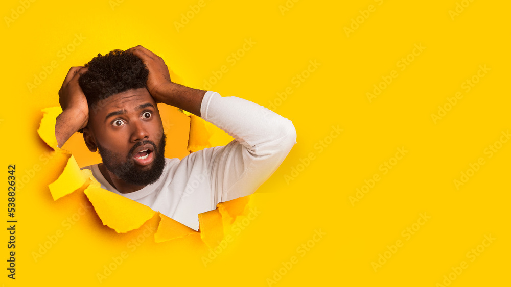 Shocked millennial african american guy holding with hands his head and looking through hole in yellow paper Shocked millennial african american guy holding with hands his head and looking through hole in yellow paper