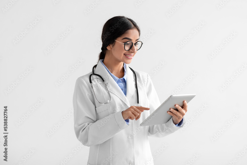Happy young indian lady doctor in white coat and glasses working on tablet Happy young indian lady doctor in white coat and glasses working on tablet