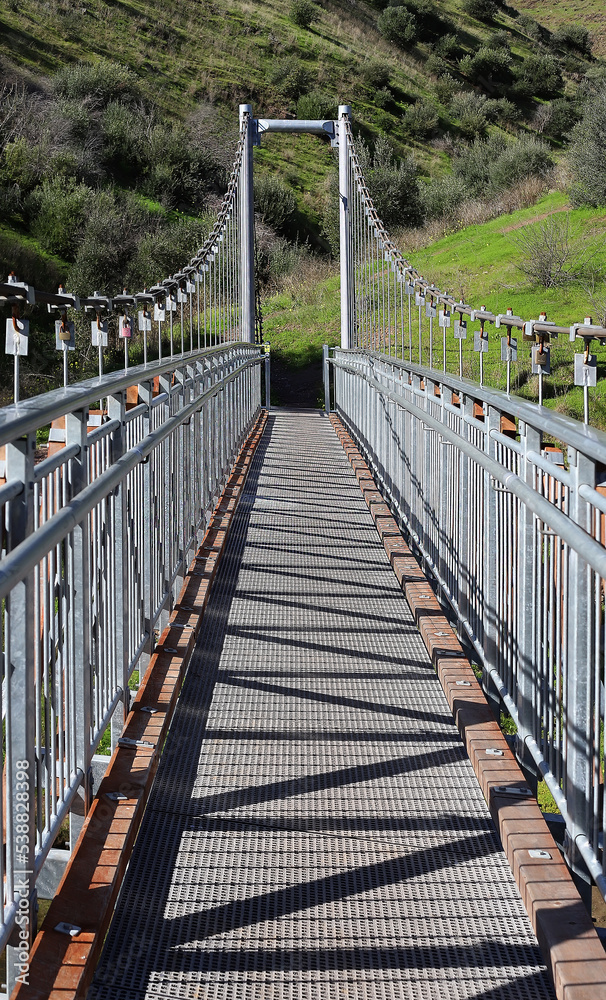 Suspension metal bridge across the river with a green hill in the background, interesting geometric picture, vertical view