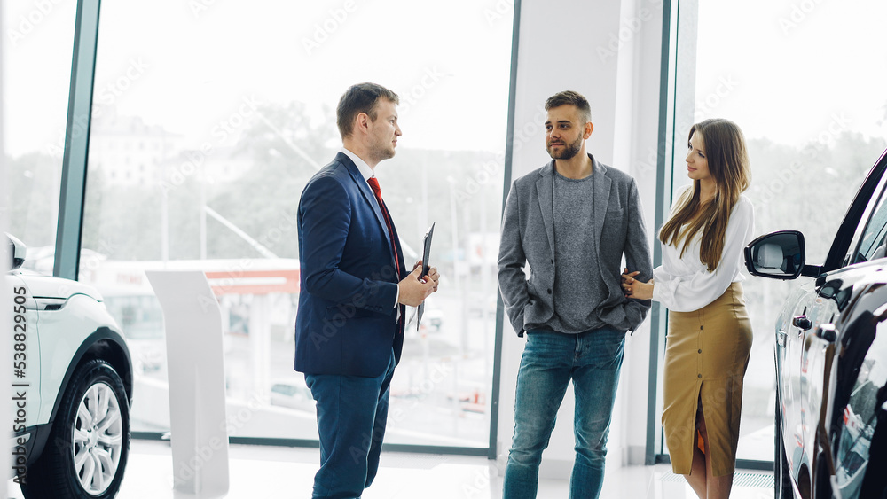 Professional car salesman is telling interested buyers beautiful couple about luxurious car in motor show while man and woman are looking at auto and listening to polite dealer. Professional car salesman is telling interested buyers beautiful couple about luxurious car in motor show while man and woman are looking at auto and listening to polite dealer.