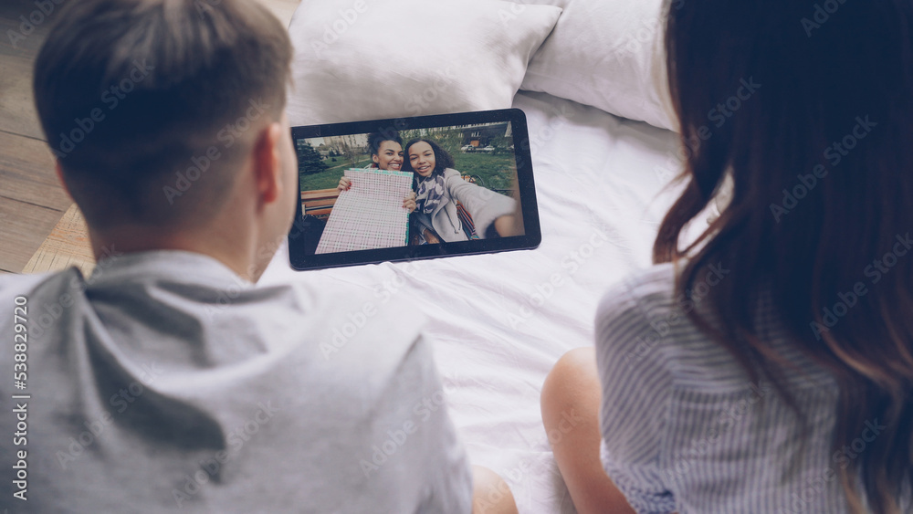 Husband and wife are talking to female friends online making video call via tablet, cheerful women on screen are showing shopping bags. Modern technology and communication concept. Husband and wife are talking to female friends online making video call via tablet, cheerful women on screen are showing shopping bags. Modern technology and communication concept.