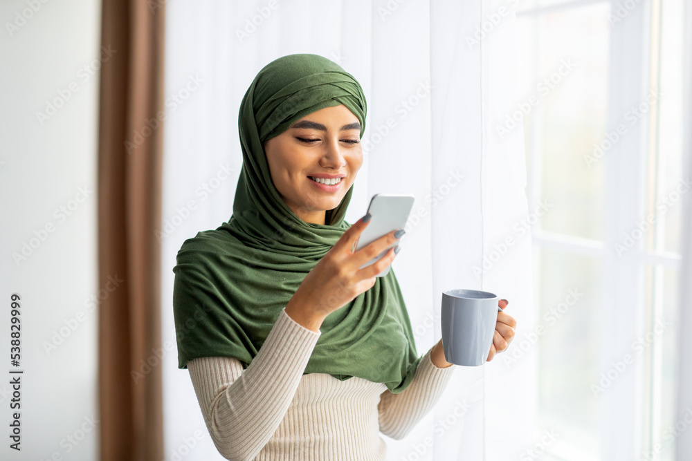 Happy young Arab woman in hijab standing near window with coffee and smartphone, browsing internet, chatting online Happy young Arab woman in hijab standing near window with coffee and smartphone, browsing internet, chatting online