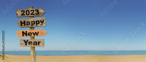 2023 happy new year written on a direction sign in front of a beach on blue sky