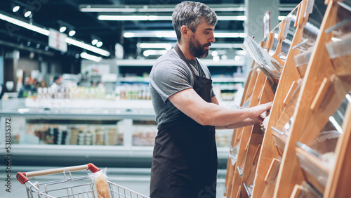 Busy male salesman is putting bread on shelves in bakery department in food store, bearded guy is wearing apron. Selling products, profession and trade concept. photo