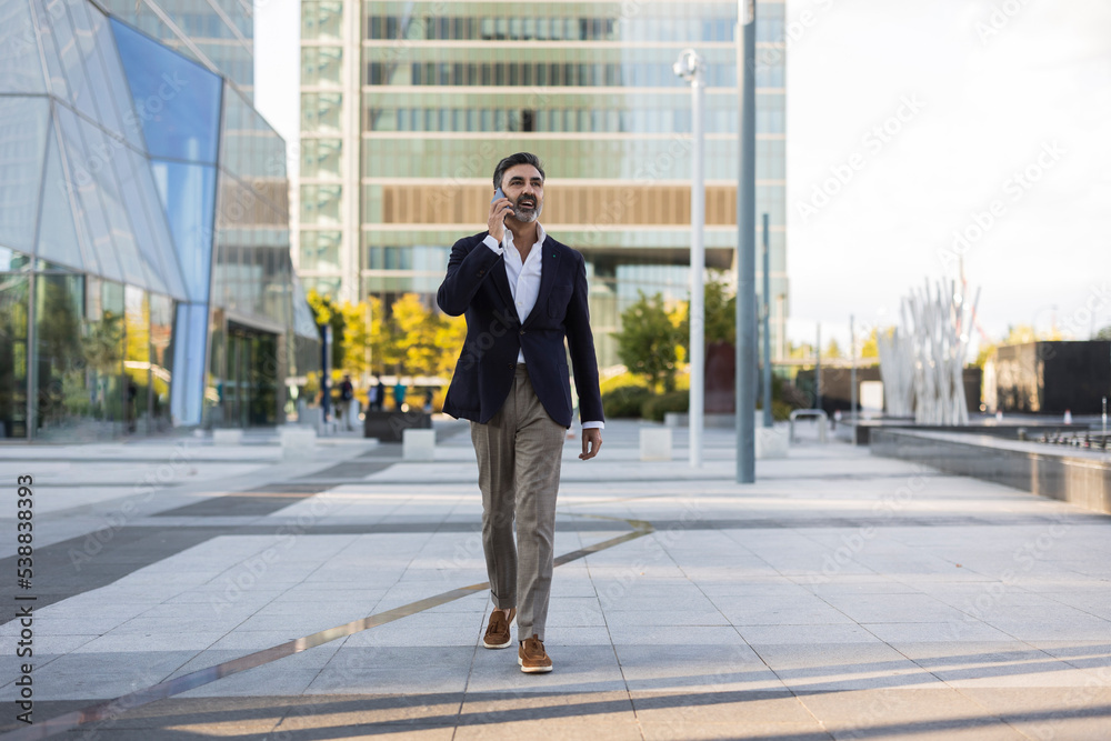 Mature businessman walking and talking on smart phone in front of office building Mature businessman walking and talking on smart phone in front of office building