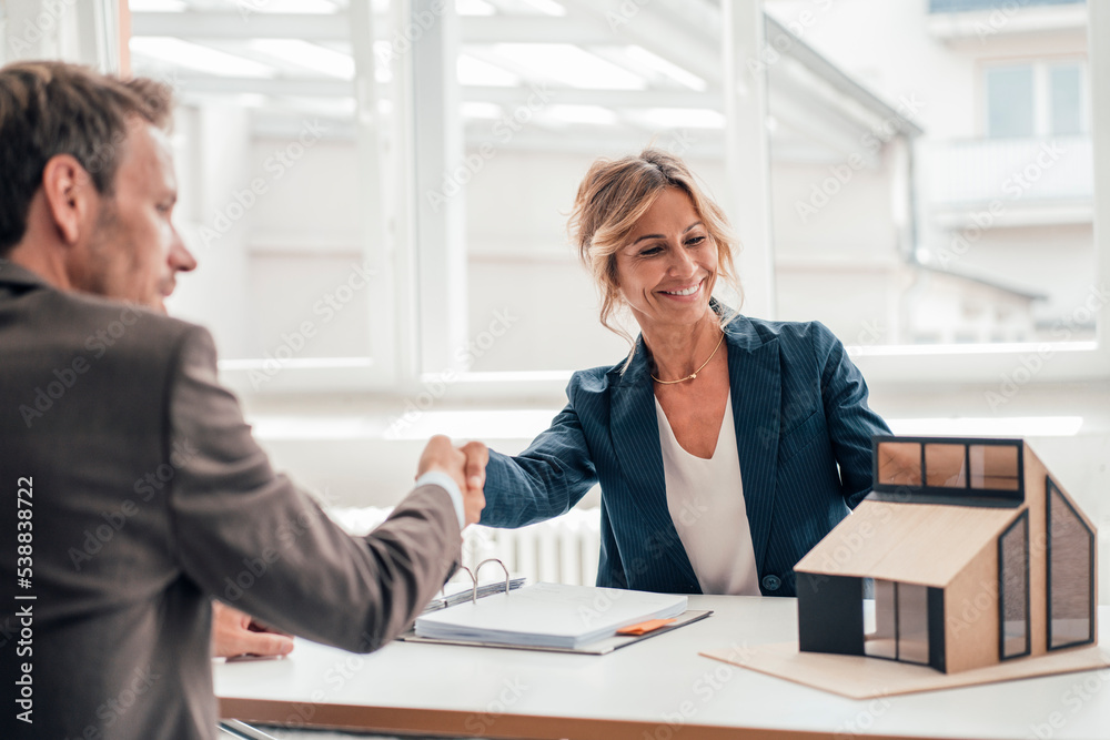 Client and real estate agent shaking hands looking at house model in office Client and real estate agent shaking hands looking at house model in office