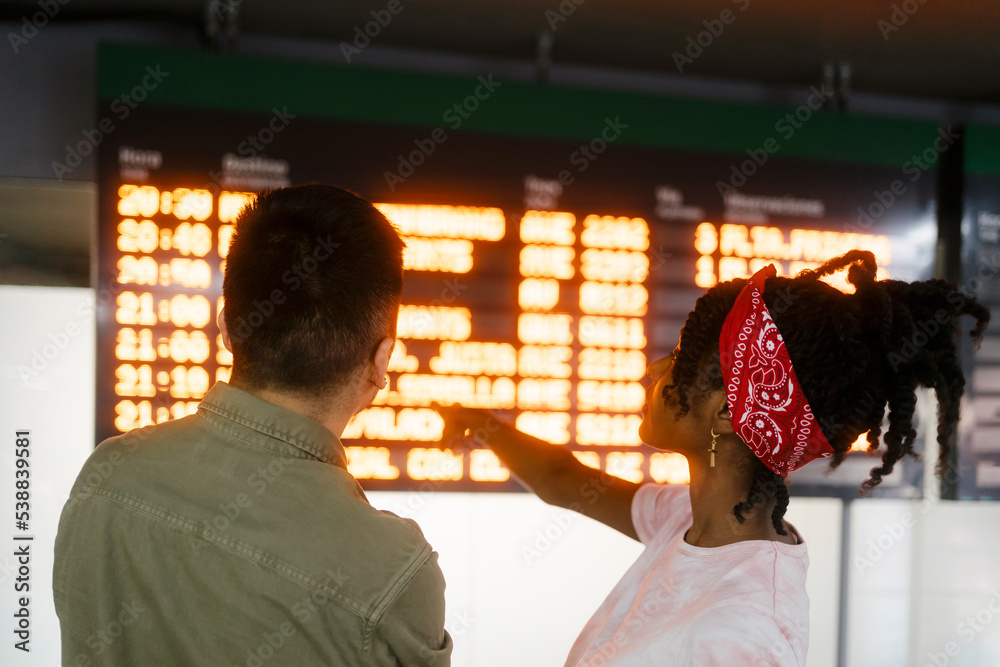 Woman with bandana pointing at departure board and talking to friend