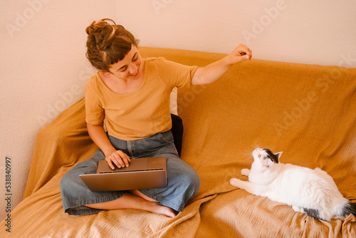 Smiling freelancer with laptop playing by cat sitting on sofa at home photo