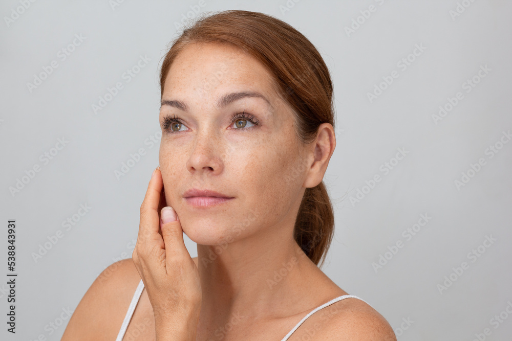 Portrait of cropped caucasian middle aged woman face with freckles holding fingers on cheek , showing soft skin on white background looking aside - obrazy, fototapety, plakaty 