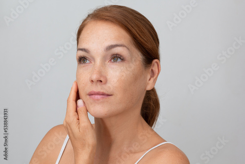 Portrait of cropped caucasian middle aged woman face with freckles holding fingers on cheek , showing soft skin on white background looking aside photo