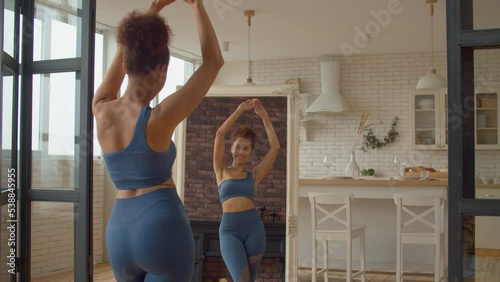 Reflection in mirror of slim smiling attractive African woman in sportswear looking at body shape, rejoicing success of dieting plan and weight loss achievement, expressing cheerful mood and happiness