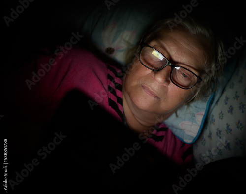 Elderly woman lies on the bed and looks into the smartphone.