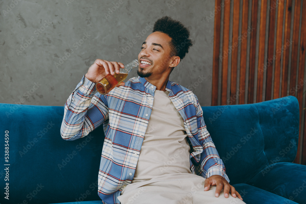 Young happy smiling fun man wear casual shirt sit on blue sofa hold bottle drink beer stay at home hotel flat rest relax spend free spare time in living room indoors grey wall. People lounge concept.