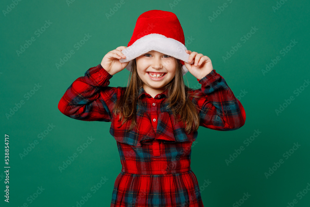 Merry smiling fun happy cute little child kid girl 6-7 years old wear red dress covering eye with Christmas hat posing look camera isolated on plain dark green background. Happy New Year 2023 concept.