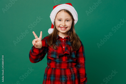 Merry smiling happy fun cool friendly little child kid girl 6-7 years old wear red dress Christmas hat posing showing victory sign isolated on plain dark green background. Happy New Year 2023 concept.