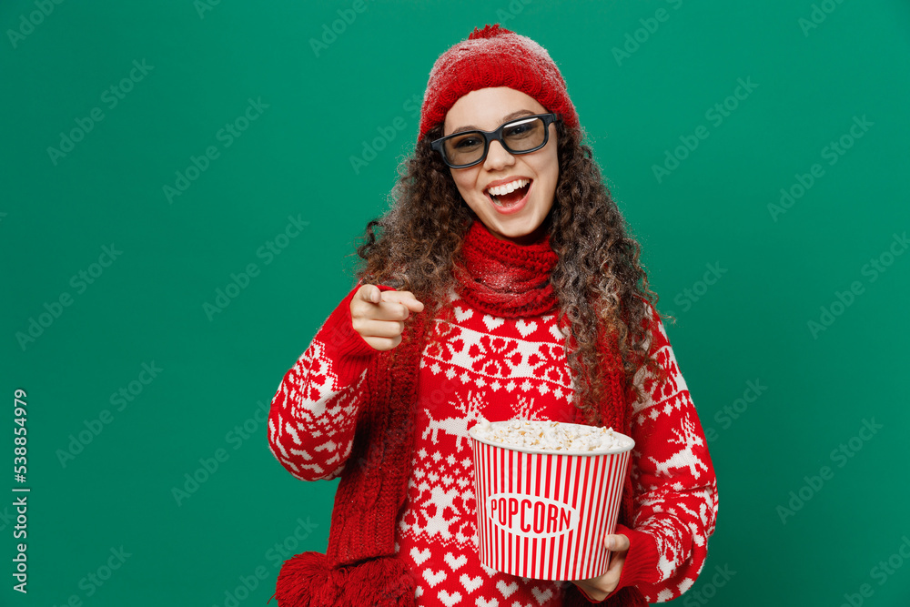 Young woman in red knitted sweater hat 3d glasses watch movie film hold bucket of popcorn in cinema point finger camera on you isolated on plain green background Happy New Year 2023 holiday concept.