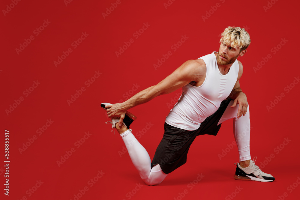 Full body young strong sporty sportsman man wear white clothes spend time in home gym train do stretch exercise for legs, sit at floor isolated on plain red background. Workout sport fit body concept.