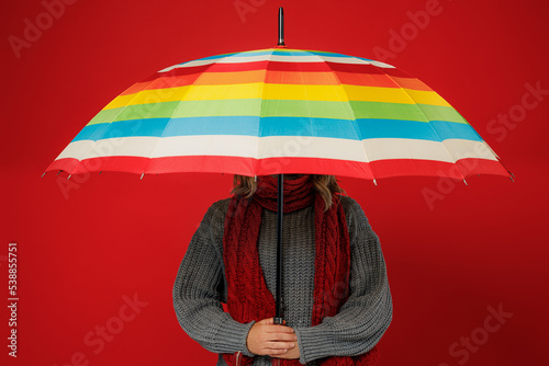 Photo Young woman wears grey sweater scarf hold cover face with opened colorful umbrella isolated on plain red background studio portrait