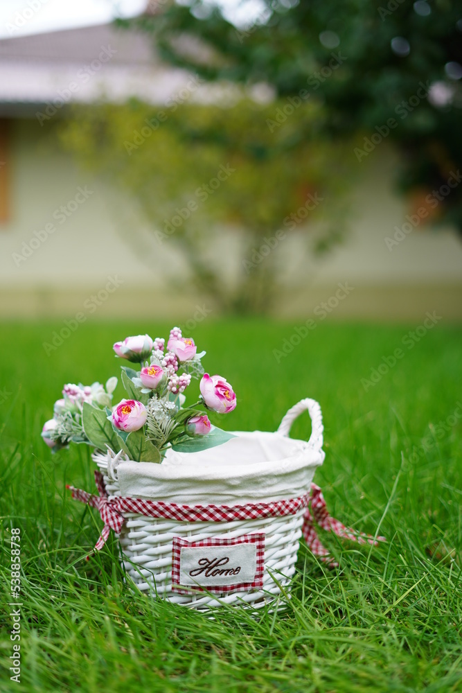 White wicker basket with decorative pink roses stands on green grass in the middle of the garden. Copy space White wicker basket with decorative pink roses stands on green grass in the middle of the garden. Copy space
