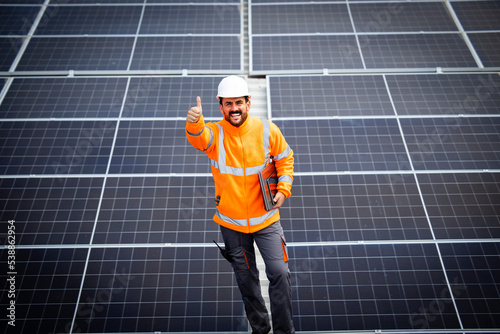 Portrait view of professional worker contractor holding thumbs up and standing in solar energy power plant after successful installation of photovoltaic cell panels.