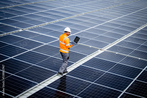 Renewable energy engineer walking through large solar power plant and checking electricity production on his laptop computer. Sustainable energy source and eco friendly technologies. photo